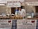 A market stall displays a variety of cheeses and other local food products. Two people are tending to the stall, with a man examining the goods and a woman standing beside him. The stall is covered with a checkered tablecloth, and several signs and banners advertise 'Porchetta da Pasqualino' and 'Salumi di Norcia'. There are stacks of cheese wheels and baskets of meats set on the table under a canopy.