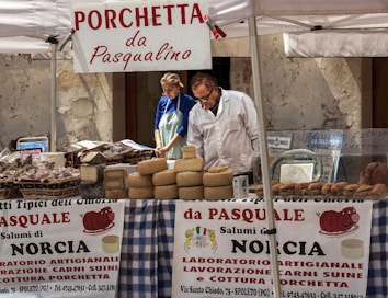 A market stall displays a variety of cheeses and other local food products. Two people are tending to the stall, with a man examining the goods and a woman standing beside him. The stall is covered with a checkered tablecloth, and several signs and banners advertise 'Porchetta da Pasqualino' and 'Salumi di Norcia'. There are stacks of cheese wheels and baskets of meats set on the table under a canopy.