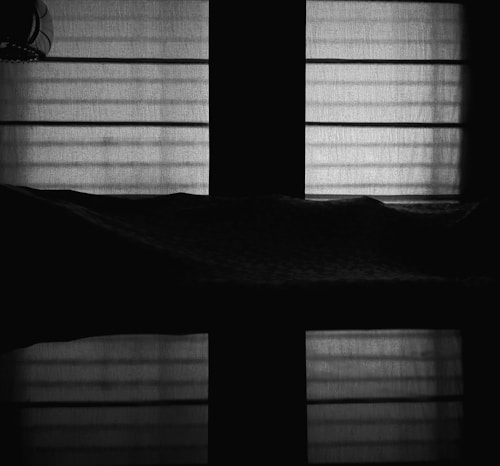 A dimly lit room features backlit window blinds casting shadows and patterns on a surface. The upper portion shows vertical and horizontal lines, while the lower part is a darker silhouette, offering minimal detail.