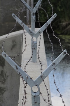 A close-up view of metal posts with barbed wire stretched across them, set along a concrete wall. The wire appears to be arranged in a zigzag pattern, and there is a body of water visible to the right side. The background is blurred with greenery.