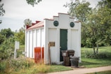 A small, white, rectangular utility building with a dark green door and red trim is situated in a grassy area surrounded by trees. Two round vents are visible on the front. In front of the building, there is a brown garbage bin and a black metal trash can. Signs and orange construction fencing are present nearby.