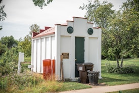 A small, white, rectangular utility building with a dark green door and red trim is situated in a grassy area surrounded by trees. Two round vents are visible on the front. In front of the building, there is a brown garbage bin and a black metal trash can. Signs and orange construction fencing are present nearby.