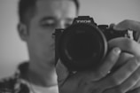 A person holding a Sony camera close to their face, with a focus on the camera lens. The image is in black and white, creating a classic and artistic feel.