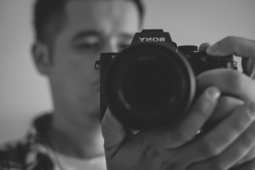 A person holding a Sony camera close to their face, with a focus on the camera lens. The image is in black and white, creating a classic and artistic feel.