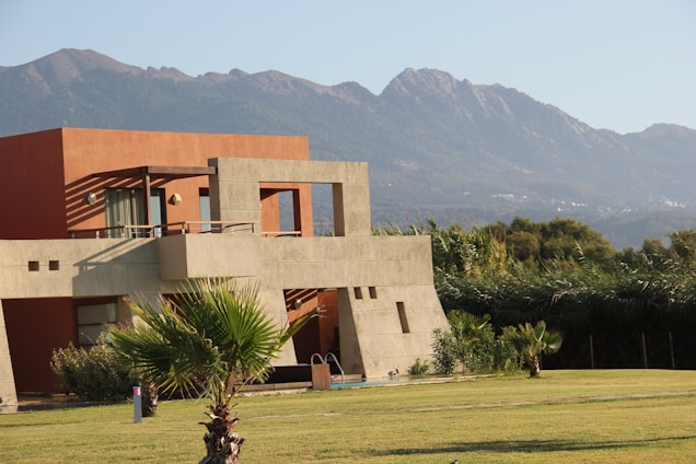 A modern house with clean architectural lines sits in front of a backdrop of green mountains. The structure features a combination of reddish-brown and beige colors, with large windows and an expansive balcony. The foreground includes a neatly manicured lawn with a few palm trees and bushes, and there is a swimming pool adjacent to the house.