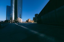 A cityscape with modern skyscrapers and tall buildings, featuring a blue sky and shadows cast on the ground. The architecture is sleek and reflective, displaying the urban environment in a serene and minimalist way.