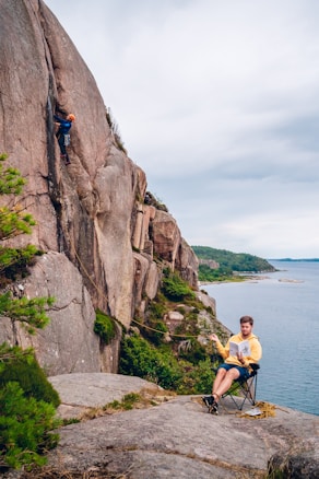 A rock climber is ascending a steep cliff face while another person sits on a camping chair nearby, holding a magazine and managing the climbing rope. The scene is set in a scenic outdoor location with lush greenery and a clear body of water in the background.