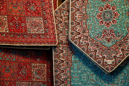 Intricately designed Persian rugs featuring detailed floral and geometric patterns. The rugs predominantly display rich red and teal colors with accents of black and beige. They are layered, showcasing different sections of the designs with ornate borders.