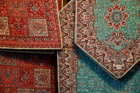 Intricately designed Persian rugs featuring detailed floral and geometric patterns. The rugs predominantly display rich red and teal colors with accents of black and beige. They are layered, showcasing different sections of the designs with ornate borders.