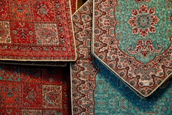 Intricately designed Persian rugs featuring detailed floral and geometric patterns. The rugs predominantly display rich red and teal colors with accents of black and beige. They are layered, showcasing different sections of the designs with ornate borders.