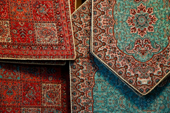 Intricately designed Persian rugs featuring detailed floral and geometric patterns. The rugs predominantly display rich red and teal colors with accents of black and beige. They are layered, showcasing different sections of the designs with ornate borders.
