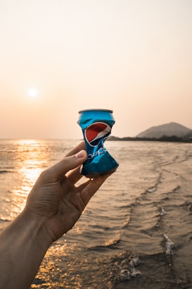 A crumpled blue can featuring a recognizable red, white, and blue logo is held up against the backdrop of a serene beach at sunset. Gentle waves cascade onto the shore beneath a golden sky, and a distant hill is visible on the horizon.