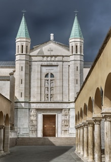 A grand stone church is flanked by two tall towers with green copper roofs, set against a dramatic dark sky. The facade is adorned with intricate sculptures and a prominent wooden door. Roman columns line the courtyard, creating an elegant pathway toward the entrance.