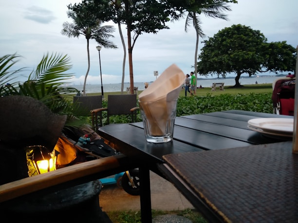 A glass holding napkins sits on a dark wooden table near a couple of empty chairs. Lush greenery, including palm trees, frames the background with a view of the ocean. A few people are visible in the distance, enjoying the beachside atmosphere. The overall setting conveys a relaxed outdoor dining experience.