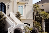A modern home facade featuring a light-colored exterior and an elegant, curved black front door with intricate glass work. A wide staircase with white railings leads up to the entrance, surrounded by manicured shrubs and palm trees. Sunlight casts shadows on the facade, highlighting the architectural details and clean lines of the house.