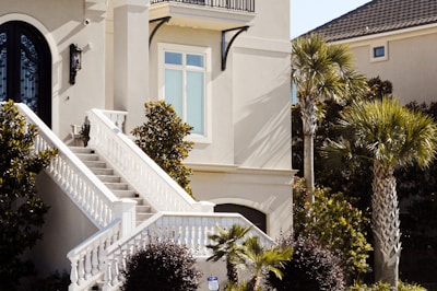 A modern home facade featuring a light-colored exterior and an elegant, curved black front door with intricate glass work. A wide staircase with white railings leads up to the entrance, surrounded by manicured shrubs and palm trees. Sunlight casts shadows on the facade, highlighting the architectural details and clean lines of the house.