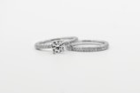Two silver rings featuring small, sparkling diamonds. The focus is on one ring with a prominent round diamond centerpiece, while the other is a plain band with embedded diamonds, arranged side by side against a white background.