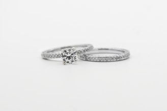 Two silver rings featuring small, sparkling diamonds. The focus is on one ring with a prominent round diamond centerpiece, while the other is a plain band with embedded diamonds, arranged side by side against a white background.