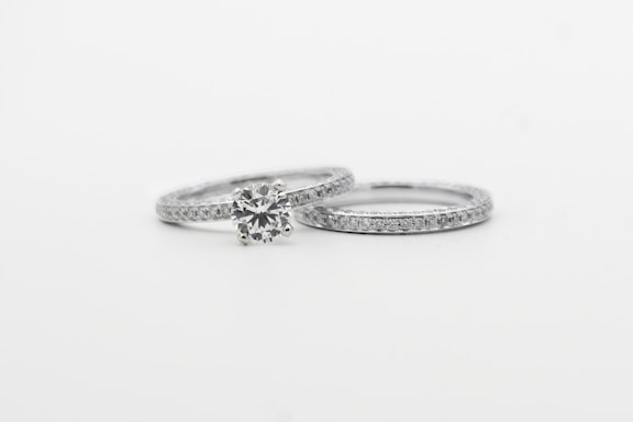 Two silver rings featuring small, sparkling diamonds. The focus is on one ring with a prominent round diamond centerpiece, while the other is a plain band with embedded diamonds, arranged side by side against a white background.