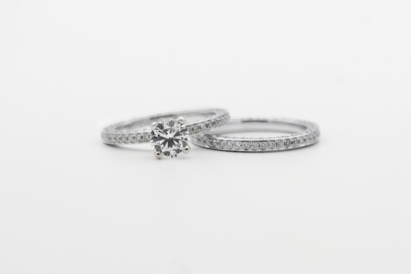 Two silver rings featuring small, sparkling diamonds. The focus is on one ring with a prominent round diamond centerpiece, while the other is a plain band with embedded diamonds, arranged side by side against a white background.