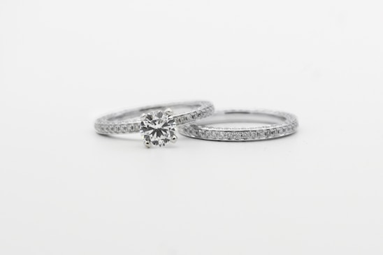 Two silver rings featuring small, sparkling diamonds. The focus is on one ring with a prominent round diamond centerpiece, while the other is a plain band with embedded diamonds, arranged side by side against a white background.