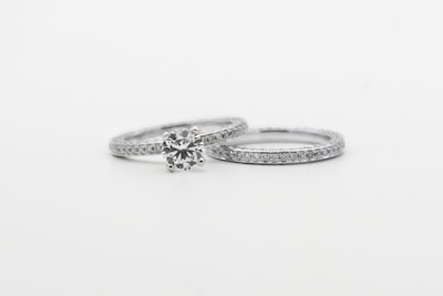 Two silver rings featuring small, sparkling diamonds. The focus is on one ring with a prominent round diamond centerpiece, while the other is a plain band with embedded diamonds, arranged side by side against a white background.