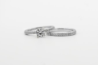 Two silver rings featuring small, sparkling diamonds. The focus is on one ring with a prominent round diamond centerpiece, while the other is a plain band with embedded diamonds, arranged side by side against a white background.