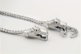A detailed image of a silver necklace featuring two intricately designed dragon heads. The dragons have red gemstones as eyes and are crafted with scales, emphasizing their fierce appearance. The chain is adorned with a series of small, sparkling stones, enhancing the luxurious feel.