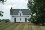 A white two-story house with a gabled roof and two chimneys is surrounded by grass and trees. The house has a central upper window and two lower windows adjacent to a front door. An outdoor grill is positioned on the left side near the house.
