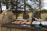 A person is grilling meat on a large outdoor stone barbecue grill surrounded by trees. There are multiple trays of food, utensils, and water bottles on the grill. The person is dressed in a cap, vest, and long-sleeved shirt.