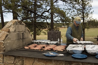 A person is grilling meat on a large outdoor stone barbecue grill surrounded by trees. There are multiple trays of food, utensils, and water bottles on the grill. The person is dressed in a cap, vest, and long-sleeved shirt.