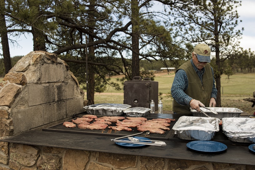 A person is grilling meat on a large outdoor stone barbecue grill surrounded by trees. There are multiple trays of food, utensils, and water bottles on the grill. The person is dressed in a cap, vest, and long-sleeved shirt.