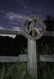 A wooden peace sign crafted from driftwood is mounted on a rustic wooden fence post, set against a backdrop of a starry night sky. A bright meteor or shooting star streaks across the sky, adding an element of wonder. Tall grass surrounds the fence, and the scene conveys a sense of tranquility and connection with nature.