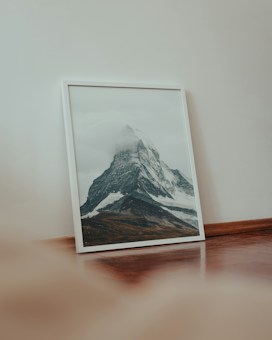 A framed photograph of a mountain with a snow-capped peak is leaning against a beige wall on a wooden floor. The mountain is partially shrouded in clouds, giving it a majestic and mystical appearance. Soft lighting and a muted color palette enhance the serene setting.