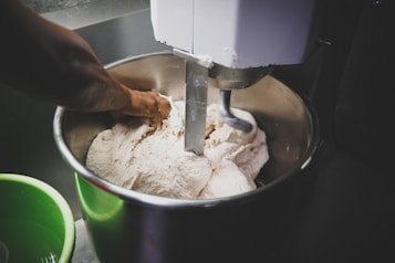 A person is kneading dough inside a large industrial mixing bowl with a mechanical dough hook attached. The scene suggests an active kitchen or bakery setting, with some green equipment visible in the foreground.