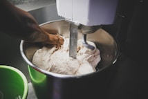 A person is kneading dough inside a large industrial mixing bowl with a mechanical dough hook attached. The scene suggests an active kitchen or bakery setting, with some green equipment visible in the foreground.