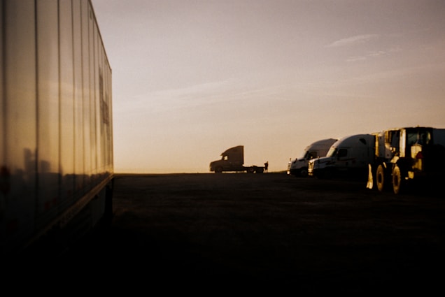 A landscape featuring several trucks parked along an open area during sunset. The silhouette of one truck stands out against the warm glow of the horizon, while other trucks and a large vehicle are aligned in a row. The light creates a soft, golden hue over the scene.