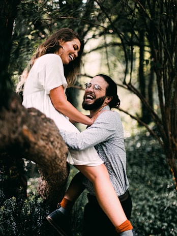 A couple is laughing and enjoying each other’s company in a forest setting. The woman, dressed in a white dress, is perched on a tree branch while the man, wearing glasses and a patterned shirt, holds her securely.
