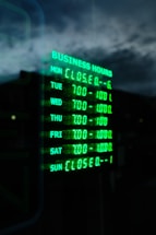 A digital sign displays business hours in glowing green text against a dark background. The days of the week are listed, with varying opening hours from Tuesday to Saturday and closures on Sunday and Monday. The sky and part of a building are faintly visible in the reflection.