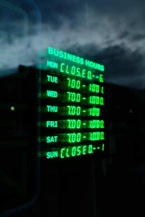 A digital sign displays business hours in glowing green text against a dark background. The days of the week are listed, with varying opening hours from Tuesday to Saturday and closures on Sunday and Monday. The sky and part of a building are faintly visible in the reflection.