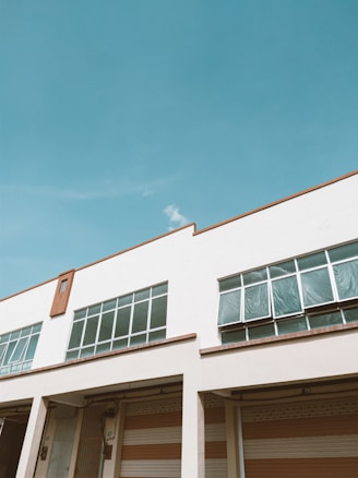 A modern industrial building with large rectangular windows and a white facade. The building has numbered garage doors below the windows. The sky is clear and blue, suggesting a sunny day.