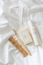 A white button-down shirt is laid out neatly with a white toothbrush, a bar of soap, a bottle of body milk, and a wooden comb arranged aesthetically on top of it. The items appear clean and organized, suggesting a minimalist and natural theme.