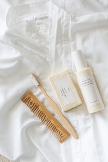 A white button-down shirt is laid out neatly with a white toothbrush, a bar of soap, a bottle of body milk, and a wooden comb arranged aesthetically on top of it. The items appear clean and organized, suggesting a minimalist and natural theme.