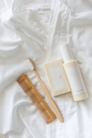 A white toothbrush with wooden handle, a brown wooden comb, a bar of soap with a minimalist label, and a bottle of body milk are neatly arranged on a soft, white fabric surface.