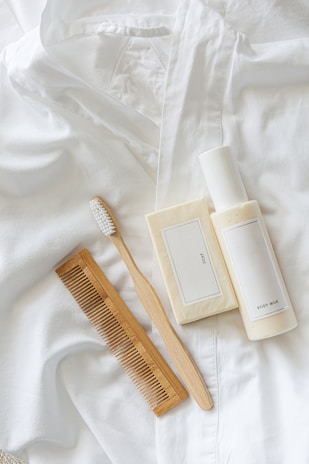 A white toothbrush with wooden handle, a brown wooden comb, a bar of soap with a minimalist label, and a bottle of body milk are neatly arranged on a soft, white fabric surface.