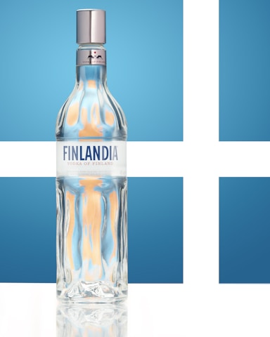 A clear glass bottle with a shiny silver cap prominently displays the label 'Finlandia Vodka of Finland' against a backdrop of stylized blue and white Nordic cross, evoking the Finnish flag.
