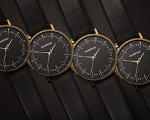 Four identical minimalist wristwatches are aligned side by side. Each watch features a black dial with simple gold markers and hands, encased in a thin gold frame. The watches are attached to sleek black leather straps, creating an elegant and cohesive appearance.
