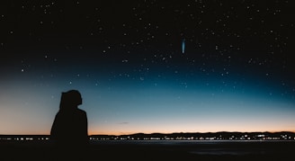 A silhouette of a person stands against a backdrop of a starry night sky. A comet with a bright tail is visible in the sky, adding a sense of wonder. The horizon features a faint glow from the setting or rising sun, casting a subtle gradient from dark blue to orange.