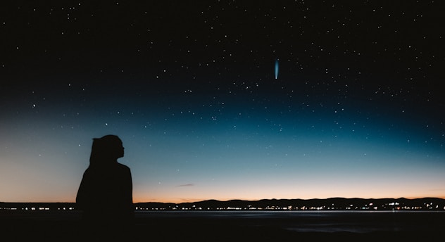 A silhouette of a person stands against a backdrop of a starry night sky. A comet with a bright tail is visible in the sky, adding a sense of wonder. The horizon features a faint glow from the setting or rising sun, casting a subtle gradient from dark blue to orange.