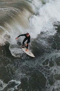 A surfer in a wetsuit skillfully maneuvers a white surfboard along a large, powerful wave. The wave is frothy and the ocean is a mix of dark green and white foamy water.